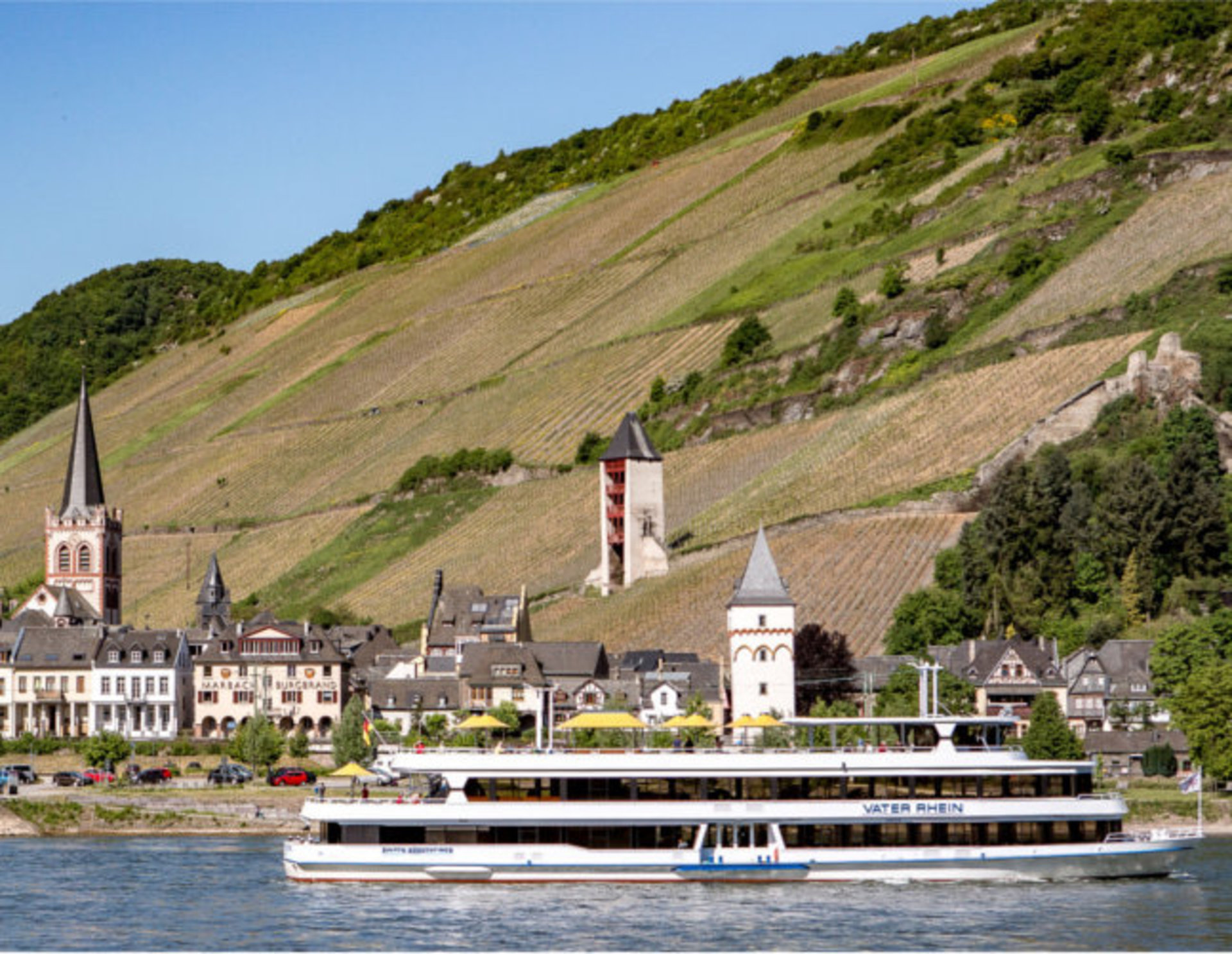 Blick auf das Rheinufer bei Rüdesheim mit historischen Fachwerkhäusern, Kirchen und Weinbergen im Hintergrund. Das Schiff "Vater Rhein" fährt auf dem Rhein. Auf dem Hügel im Hintergrund ist eine Burg zu sehen.
